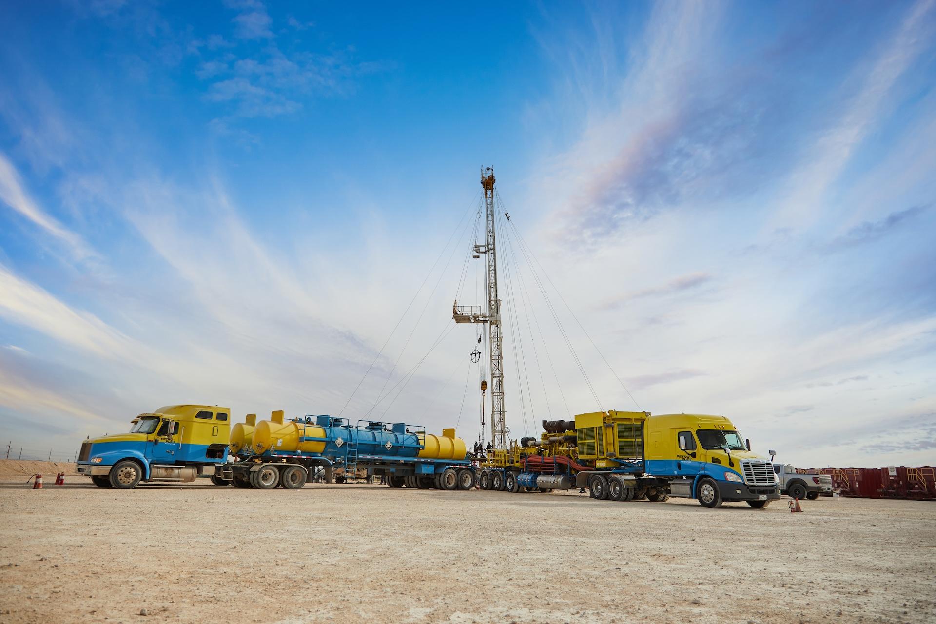 A tanker truck and a pressure-pumping truck are completing an acidizing treatment at a wellhead in the Permian Basin. The oil well services equipment is set against a bright blue sky in Midland, Texas.